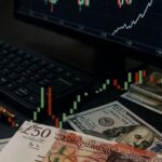 delle banconote di dollari e sterline su un piano vicino a un laptop. In sovrimpressione un grafico a candele