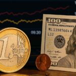 una moneta da un euro con dei grafici finanziari sullo sfondo e una banconota di una valuta diversa accanto